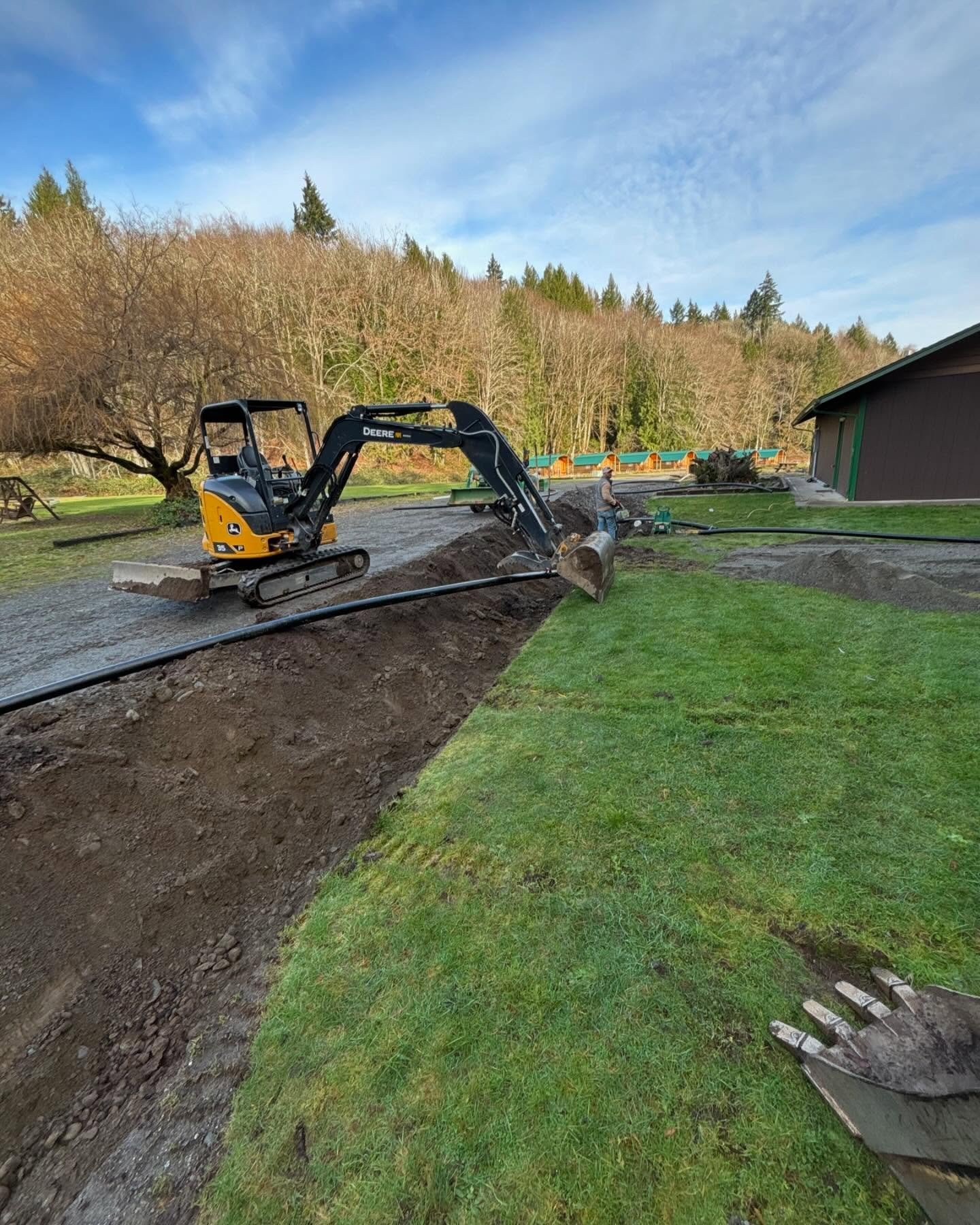 A compact excavator cutting a utility trench beside a gravel drive.
